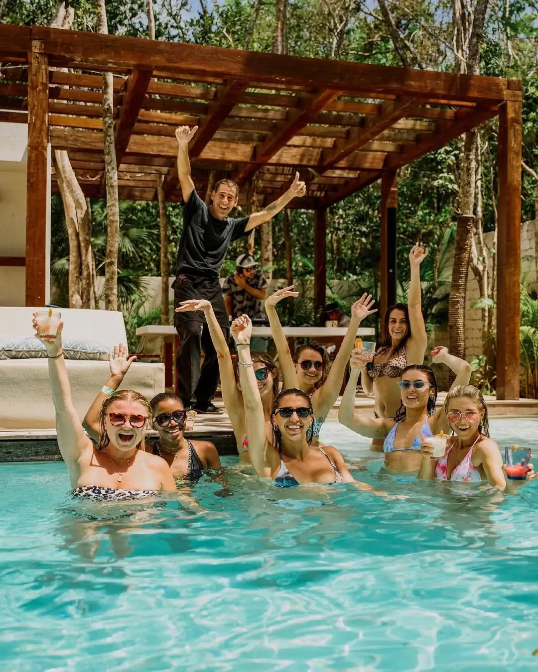 A group of guests cheering with drinks in the pool of an Aldea Kaan Tulum villa under a wooden pergola, a man on the deck raising both arms behind them