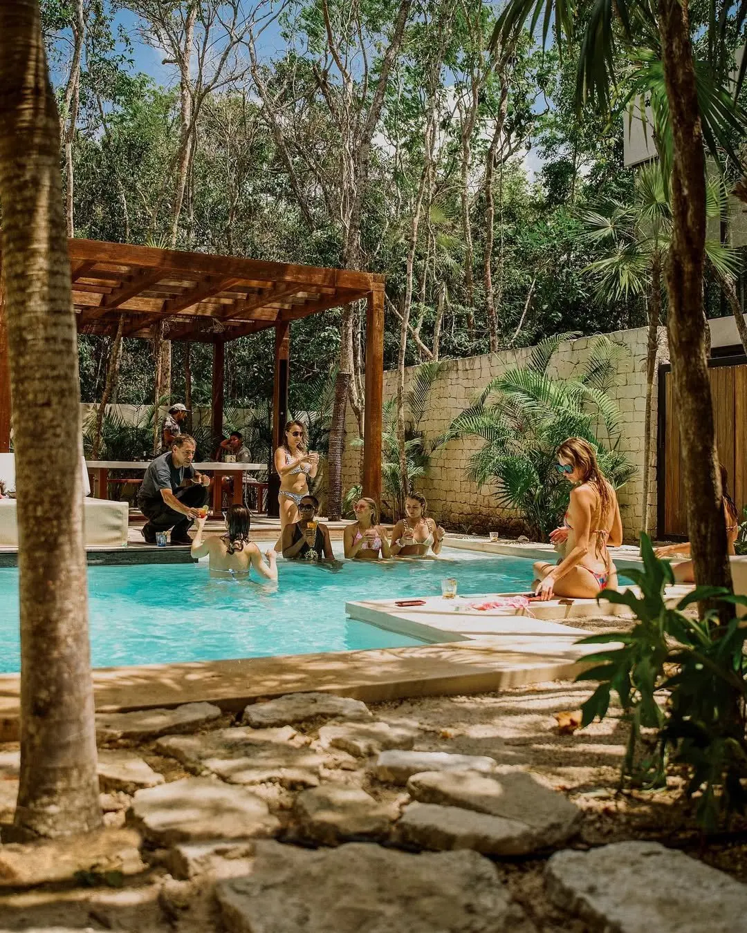 Guests with drinks in the pool of an Aldea Kaan Tulum villa beside a wooden pergola, a man on the deck handing a glass down to the water