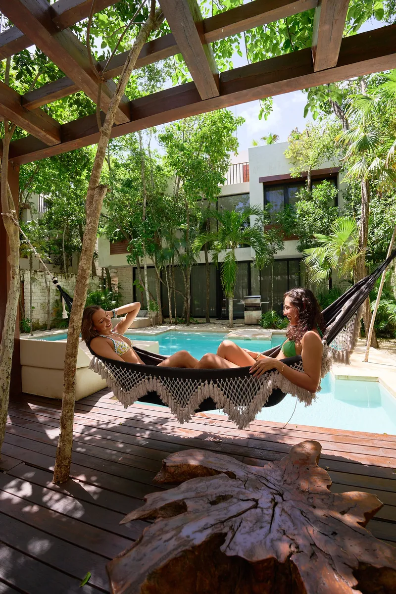Two guests in a fringed hammock under a wooden pergola beside the pool of an Aldea Kaan Tulum villa