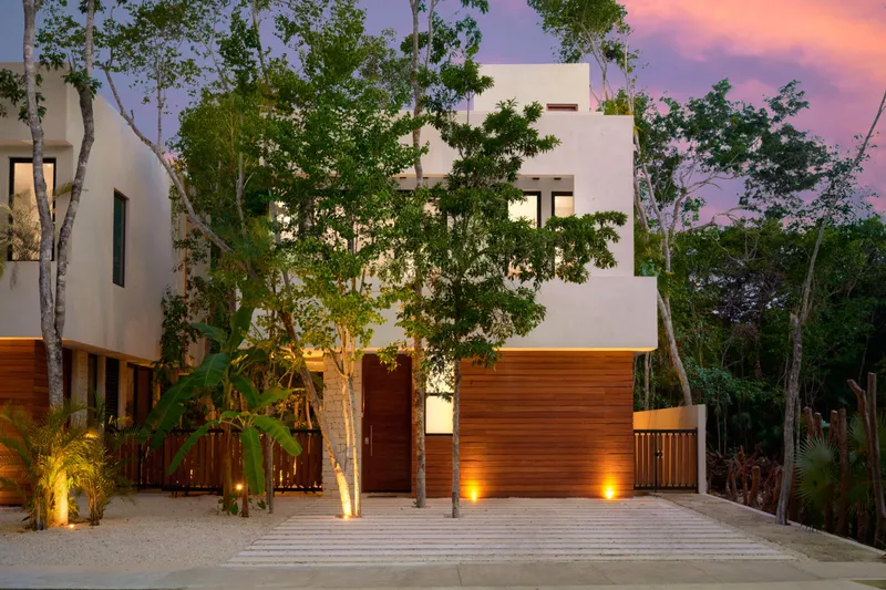 Casa David facade from the street at dusk with white upper floors, a wood clad entrance and trees in front