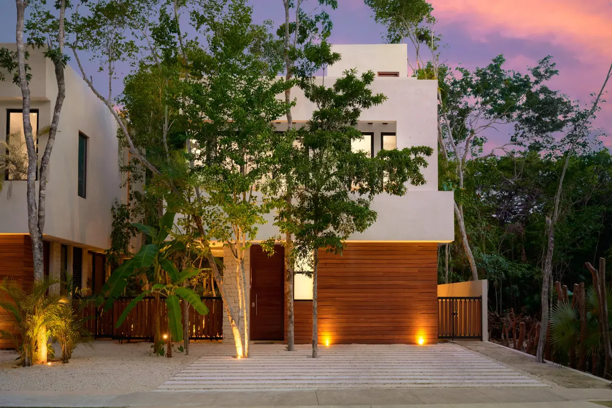 Casa David facade from the street at dusk with white upper floors, a wood clad entrance and trees in front