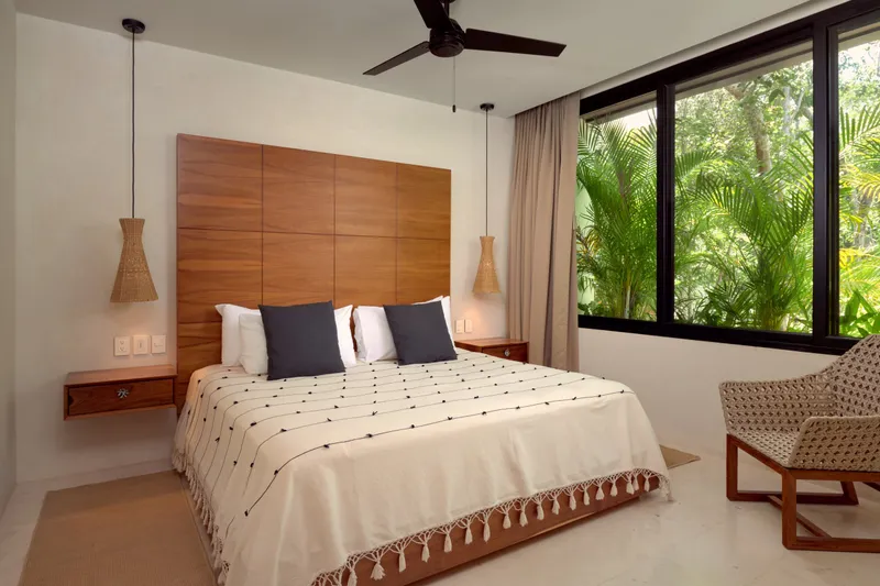 Casa Emilia bedroom with a tall wooden headboard, cream tasselled coverlet, woven pendant lamps and a window onto palms