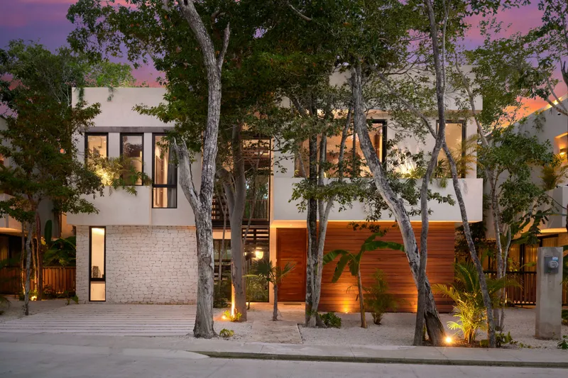 Casa Emilia facade from the street at dusk with lit windows, a wood clad entrance and tall trees