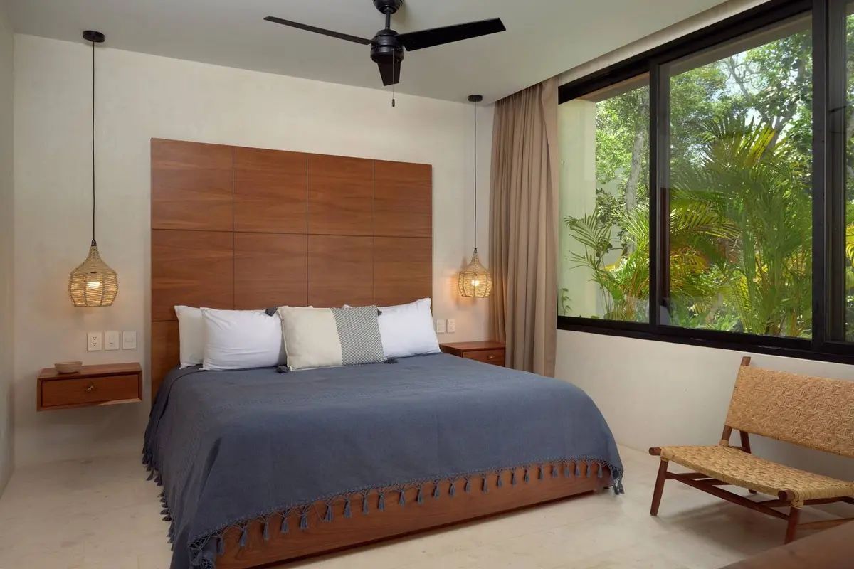Casa Lydia guest bedroom with a tall wooden headboard, blue coverlet, woven pendant lamps and a window onto palms