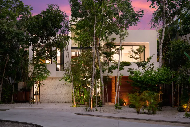 Casa Lydia facade from the street at dusk with a stone wall, a wood clad entrance and tall trees
