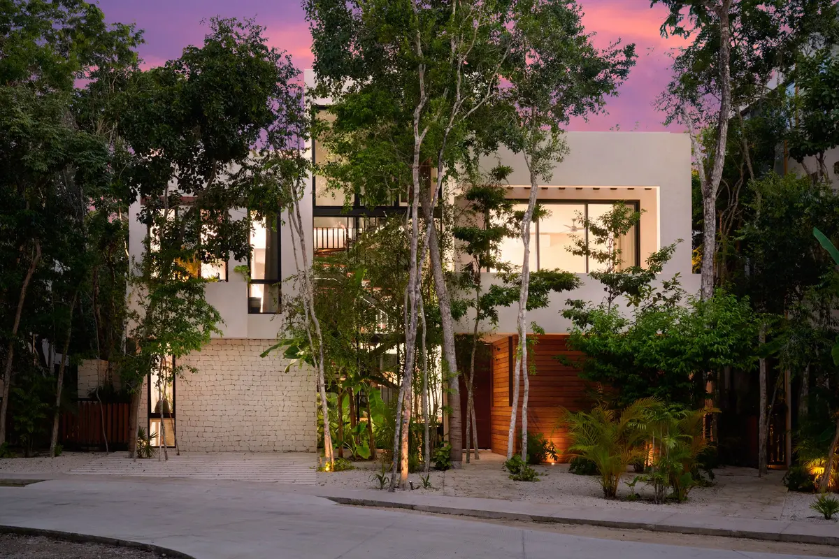 Casa Lydia facade from the street at dusk with a stone wall, a wood clad entrance and tall trees