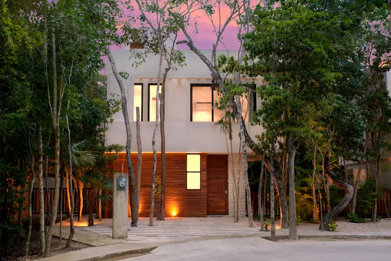 Casa Thomas facade from the street at dusk with lit windows, a wood clad ground floor and tall trees