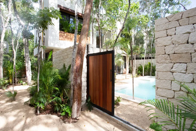 The garden gate between two Aldea Kaan Tulum villas, open: a wooden gate in a stone wall under the trees, with the pool of the villa next door beyond it