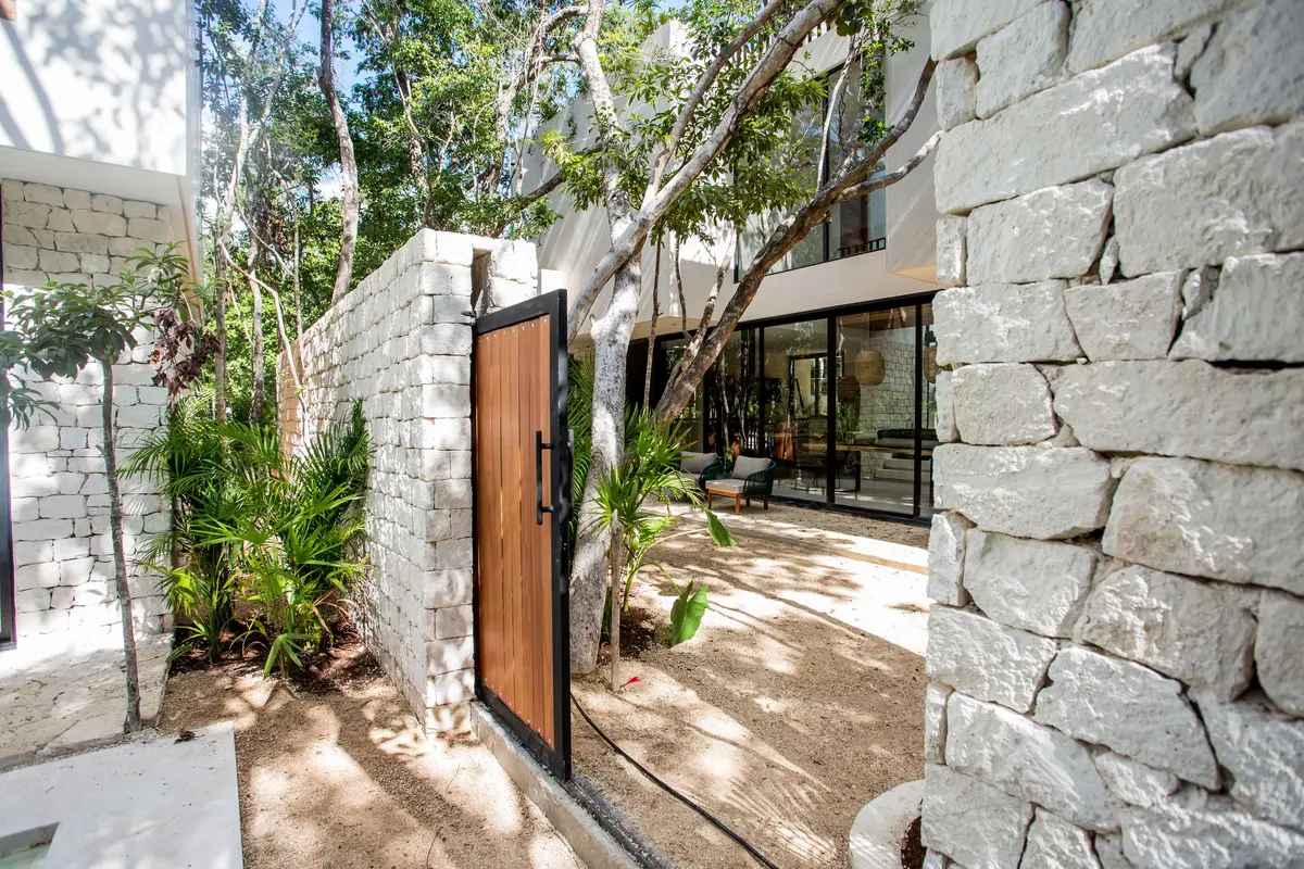The garden gate between two Aldea Kaan Tulum villas, open: a wooden gate in a white stone wall, with the glass doors and the patio of the villa next door beyond it
