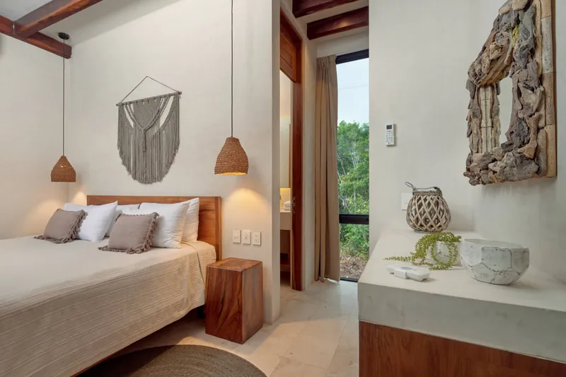 The king bed on the loft mezzanine, under woven pendant lamps and a macrame wall hanging, beside a tall window onto the jungle and an open door to the bathroom