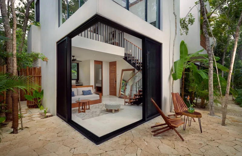 The two story loft of a rooftop pool villa at Aldea Kaan Tulum, seen from the garden, with its corner glass doors open onto the living area and two wooden folding chairs on the stone patio