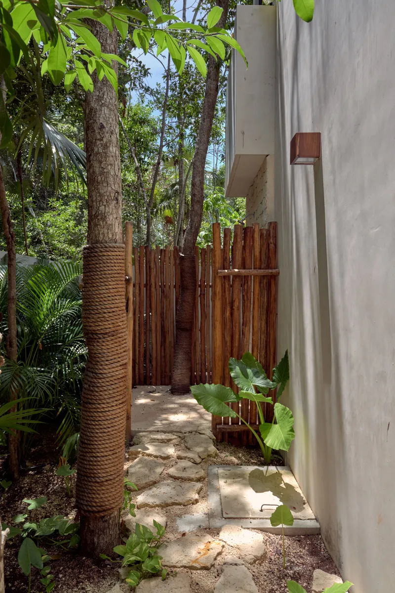 The outdoor jungle shower, a corner screened by a fence of wooden poles at the end of a stepping stone path, between a tree trunk wrapped in rope and a white wall