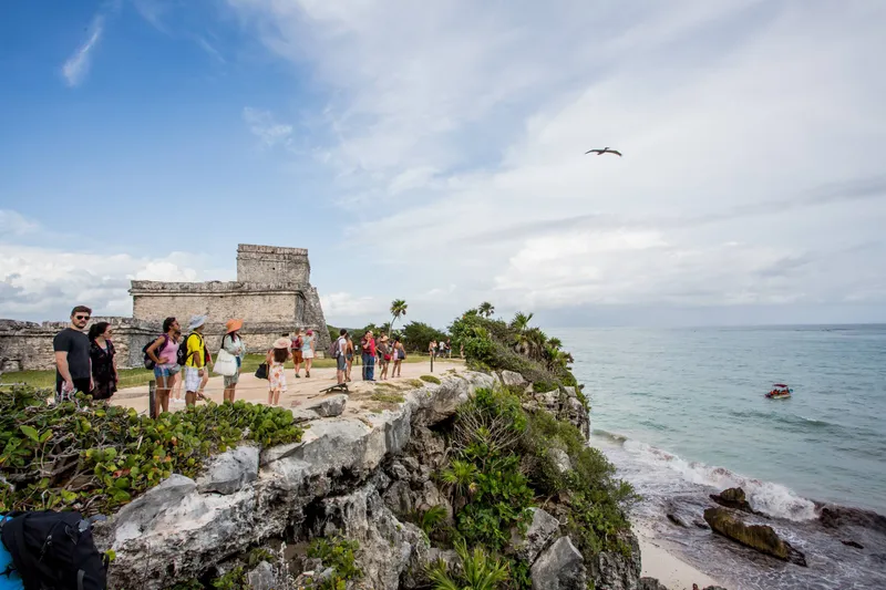 The Tulum ruins: a Mayan temple of grey stone on the cliff above the Caribbean Sea, with visitors on the path and a small boat offshore