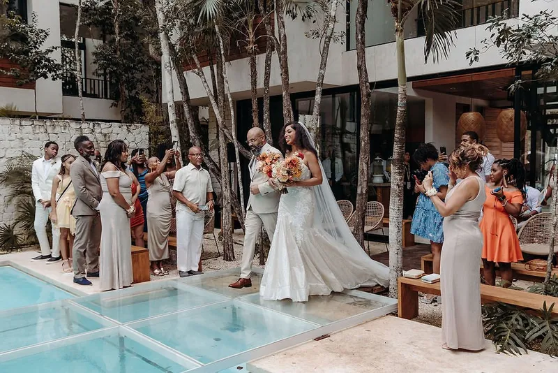 A bride on her way to the ceremony at Aldea Kaan Tulum, crossing a glass floor laid over the pool, with guests standing on both sides