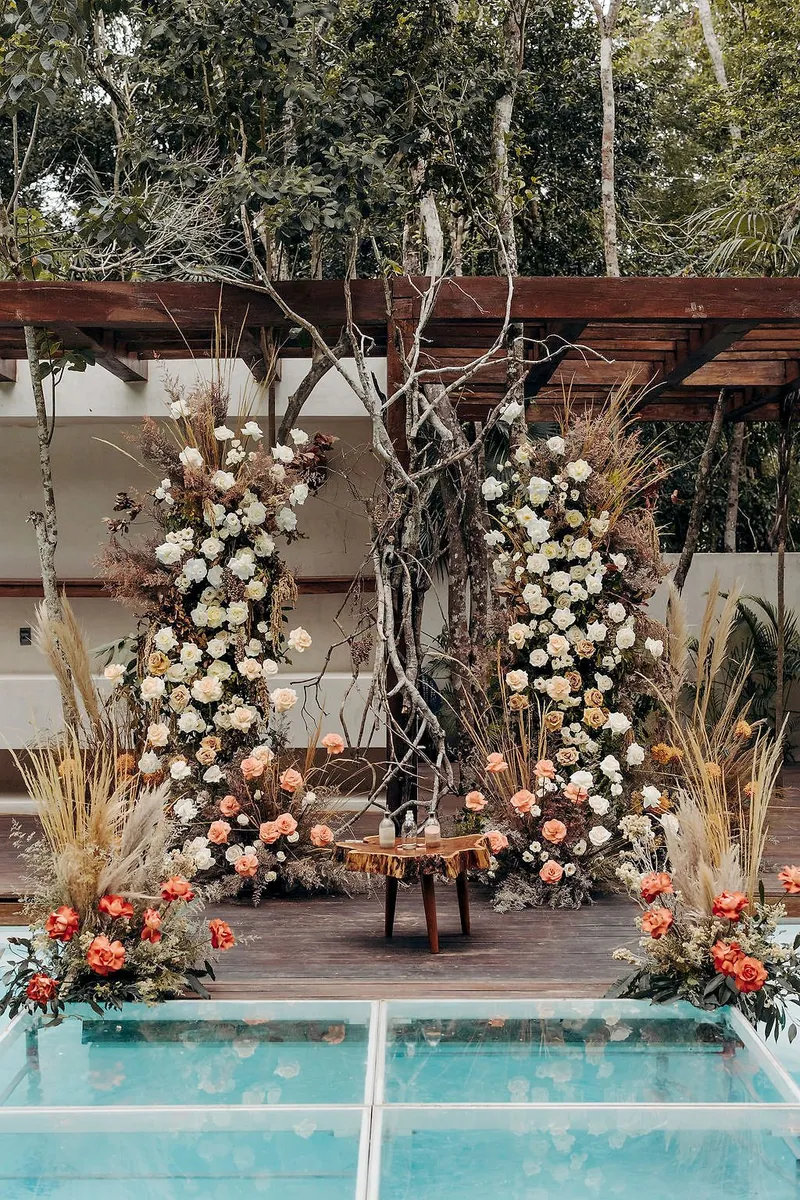 A wedding ceremony set up at Aldea Kaan Tulum before the guests arrive: two pillars of white, peach and rust flowers on a wooden deck under a pergola, with a glass walkway over the pool in front