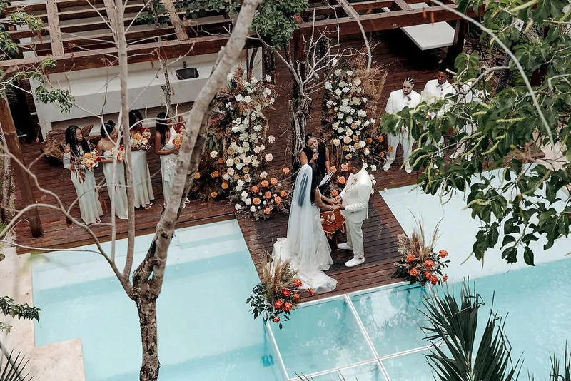 A wedding ceremony at Aldea Kaan Tulum seen from above: the couple at a flower arch on a wooden deck beside a pool, with the bridal party on one side