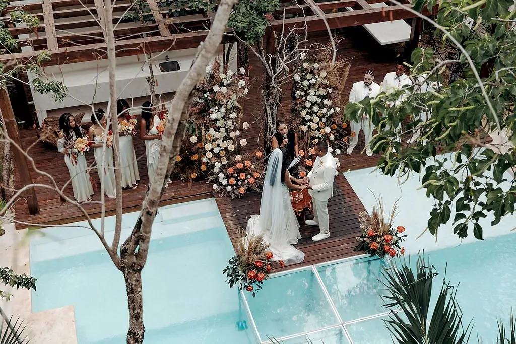A wedding ceremony at Aldea Kaan Tulum seen from above: the couple at a flower arch on a wooden deck beside a pool, with the bridal party on one side