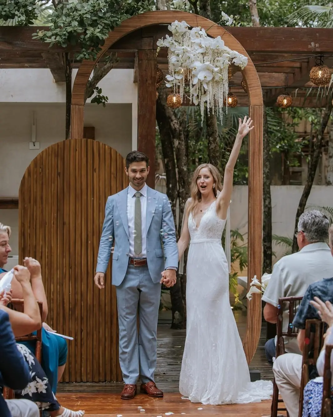 A couple after their wedding ceremony at Aldea Kaan Tulum: a wooden arch hung with white orchids behind them, bubbles in the air and guests seated on both sides