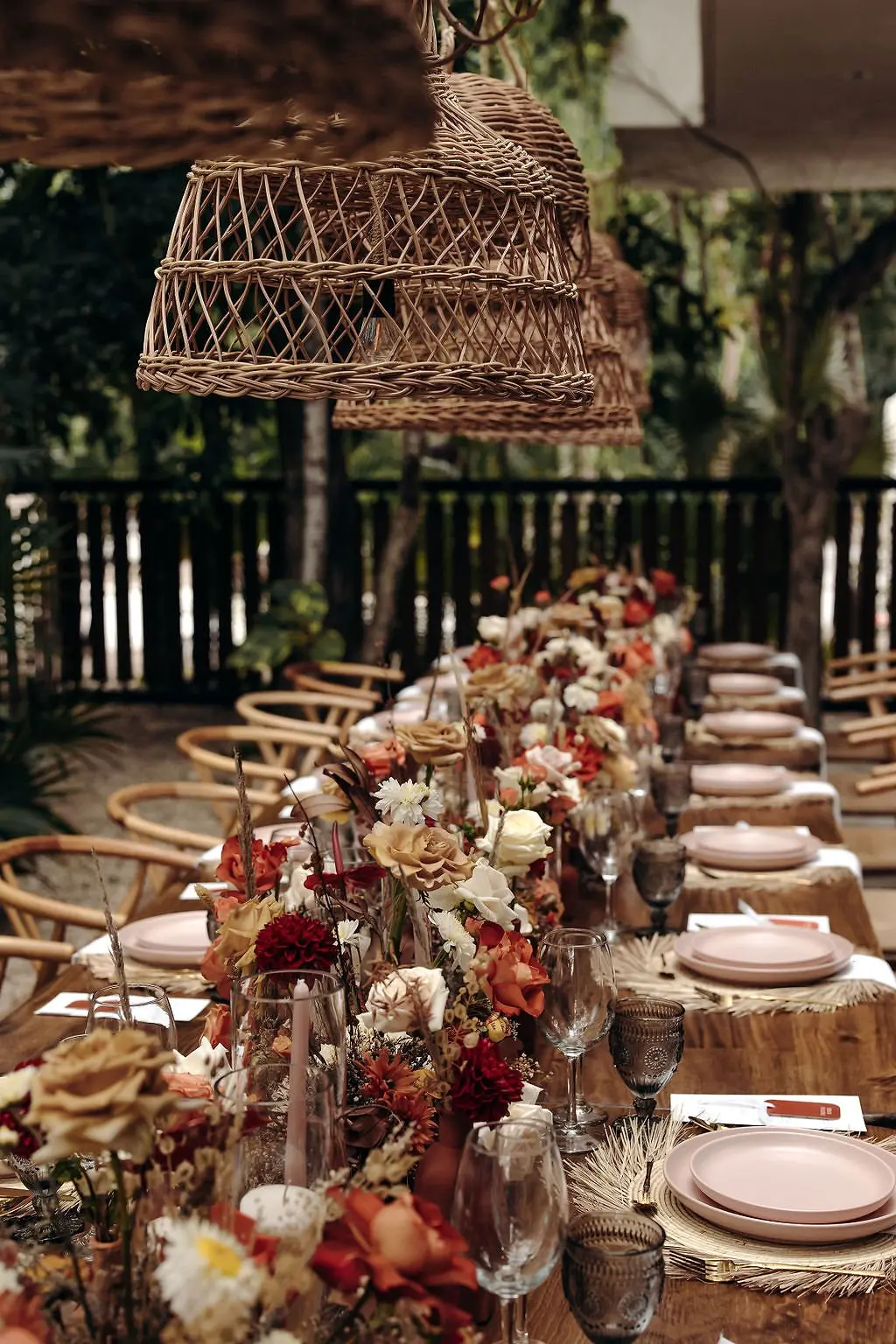 A wedding dinner table at Aldea Kaan Tulum: rattan pendant lamps over a long wooden table set with rust, peach and white flowers and pink plates