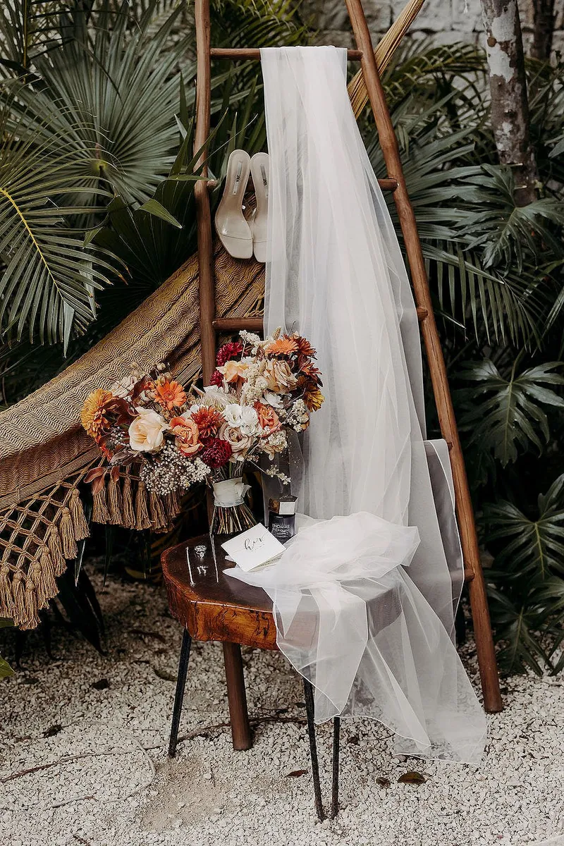 A wedding veil and shoes on a wooden ladder at Aldea Kaan Tulum, beside a bouquet of orange, rust and white flowers on a stump table